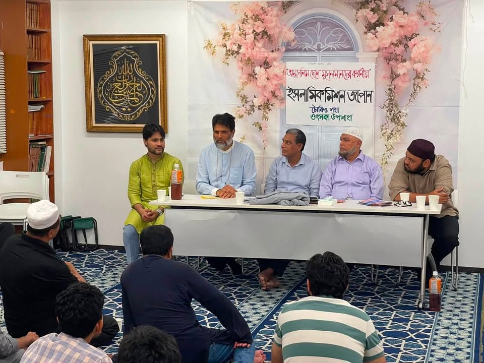 Five men sit behind a table at the front of a room with a banner reading "Islamic Mission Japan, Tokyo, Otsuka Sub-Chapter" in Bengali, while a group of attendees sit on the carpeted floor listening.