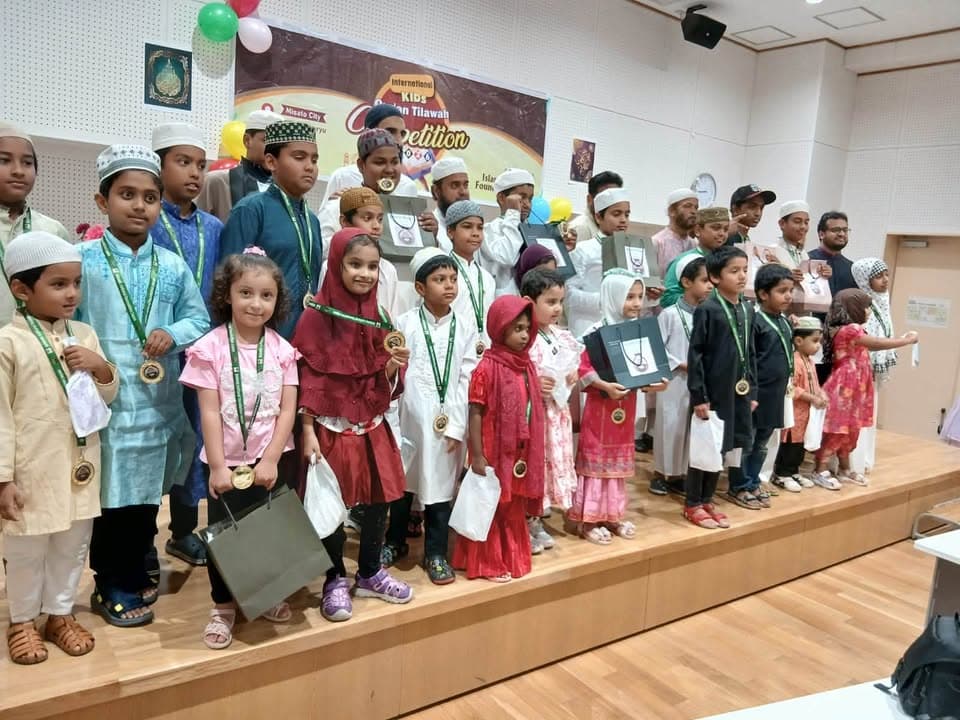 A large group of children in traditional Islamic dress stand on a stage, many wearing medals around their necks, in front of a banner reading "International Kids Qur'an Tilawah Competition" decorated with balloons.