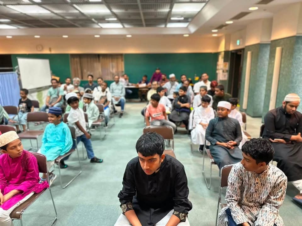 Rows of boys and men in traditional attire and prayer caps sit on chairs in a large hall, waiting attentively during an indoor gathering.