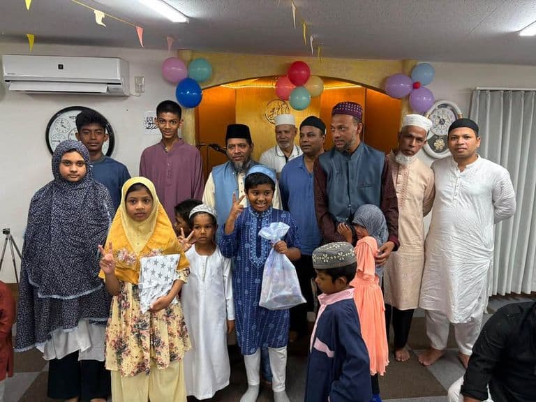 A group of men, women, and children in traditional Islamic clothing pose together for a photo inside a decorated mosque hall, with colorful balloons and streamers hanging near a golden mihrab, and some of the children holding gift bags.