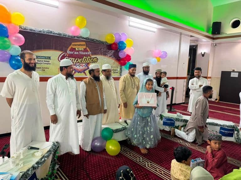 A young girl in a headscarf holds a gift box while standing among a group of men in traditional white and beige robes at a decorated mosque hall, with a colorful "International Kids Qur'an Tilawah Competition" banner and balloons in the background.