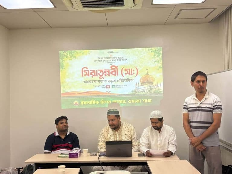 In a meeting room, three men sit at a table with a laptop while a fourth man stands speaking, in front of a projected screen displaying a Bengali-language banner for a Seerat-un-Nabi program organized by Islamic Mission Japan's Osaka chapter.