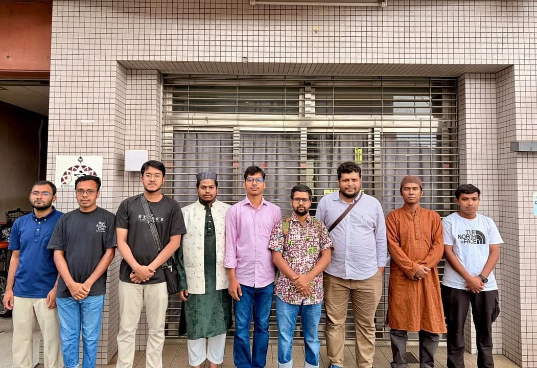 Nine men in casual and traditional clothing stand in a row outdoors, posing for a group photo in front of a building with a shuttered storefront and a Kyoto community center sign.