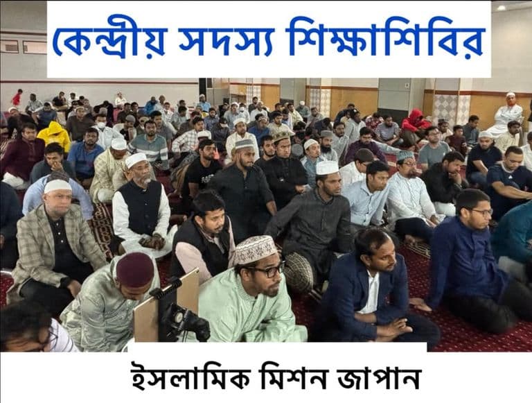 A large group of men in traditional caps and clothing sit cross-legged in rows on a carpeted hall floor, listening attentively during an indoor gathering, with a video camera set up in the foreground.