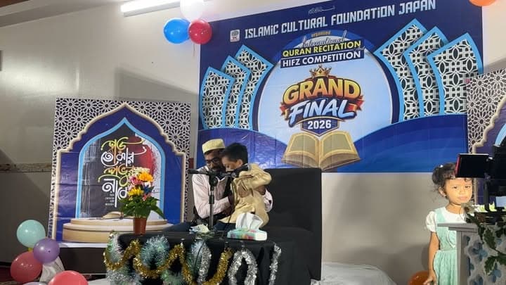 A man holding a microphone helps a young boy recite on stage in front of a large "Grand Final 2026" backdrop for the International Quran Recitation and Hifz Competition, with balloons and a decorated table nearby, while a young girl stands to the side.