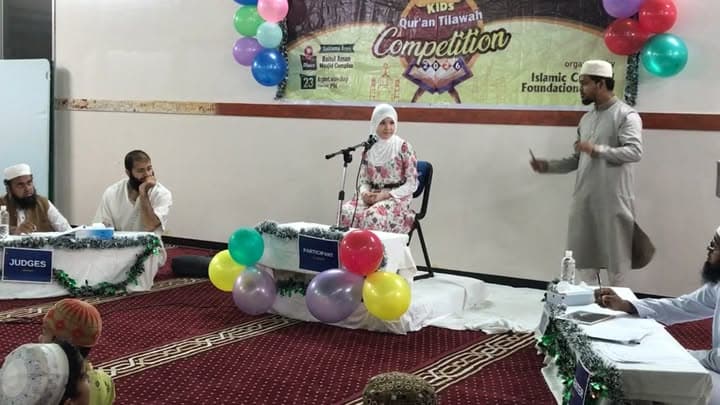 A girl in a headscarf and floral dress stands at a microphone to recite in front of judges seated at a table, with a "Kids Qur'an Tilawah Competition" banner and colorful balloons decorating the hall.